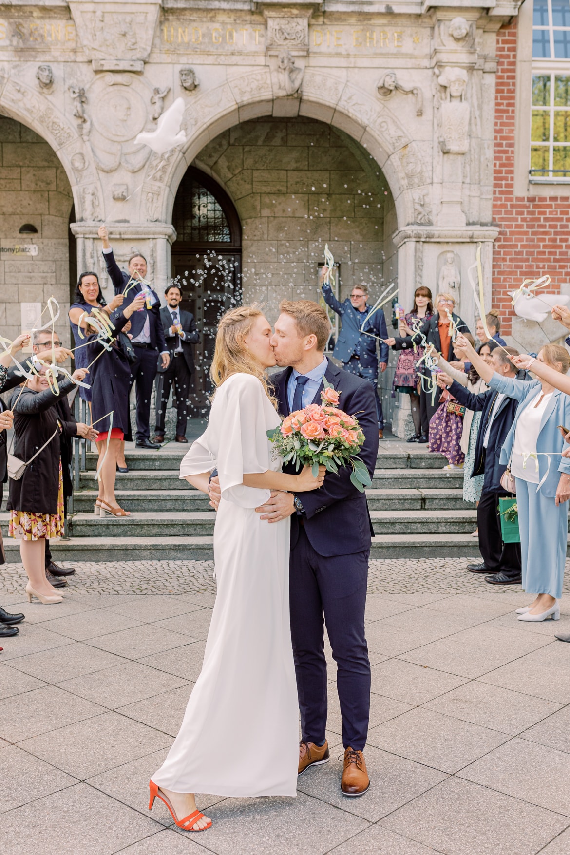 Hochzeitspaar küsst sich vor dem Standesamt während Gäste jubeln