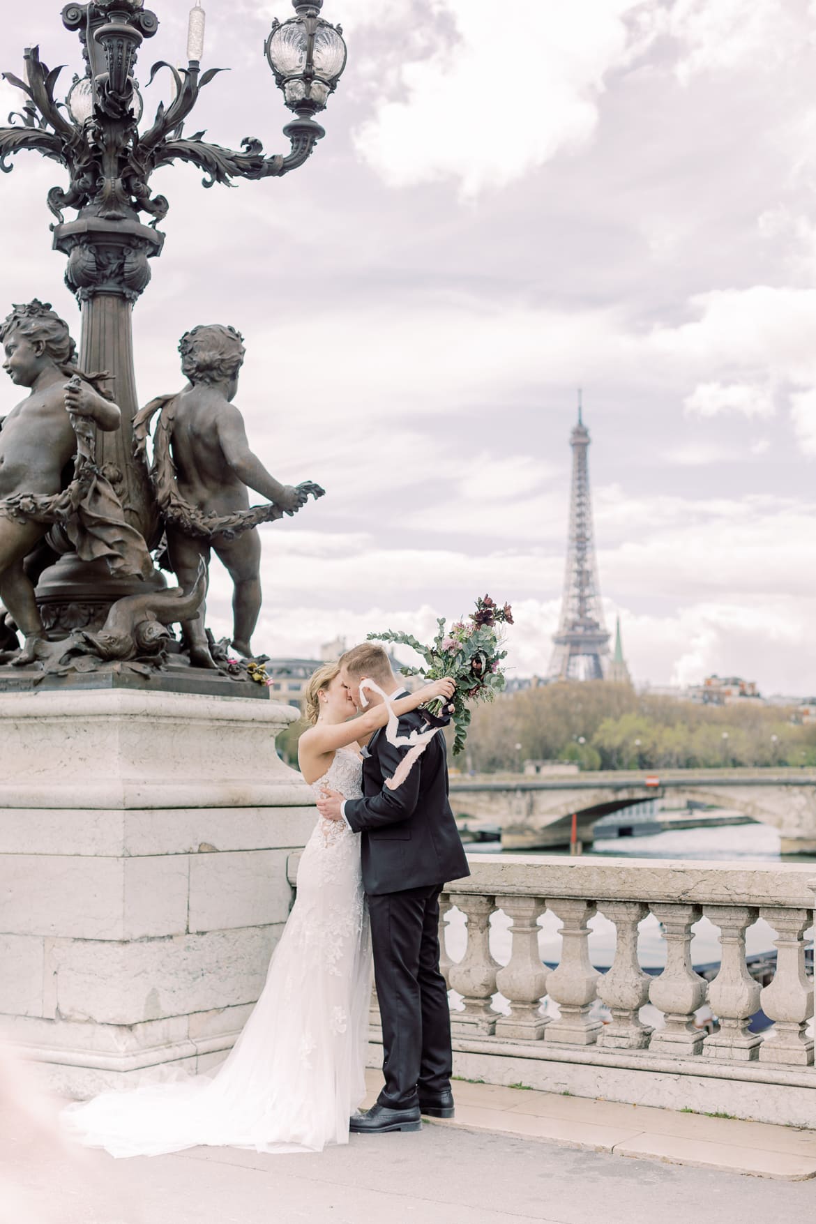 Ein Hochzeitspaar küsst sich auf einer Brücke, im Hintergrund sieht man den Eiffelturm