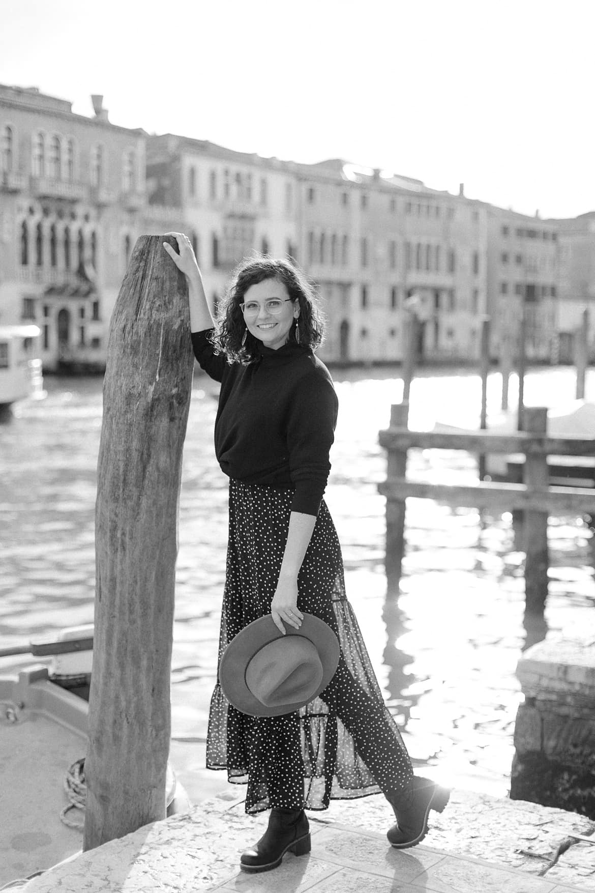 Eine Frau im schwarzen Kleid und schwarzen Pulli lehnt sich in Venedig an einen Holzpfahl am Kanal. Sie hat einen Hut in der Hand und lächelt in die Kamera.