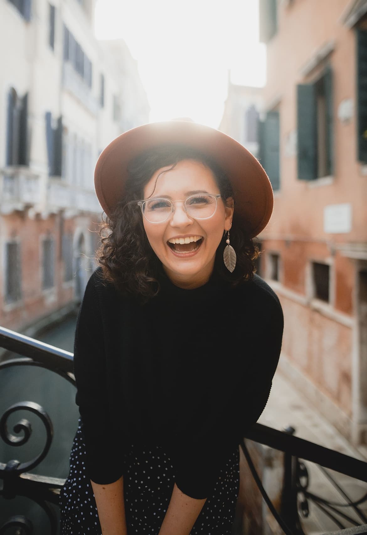 Frau mit braunen, lockigen Haaren lacht in die Kamera. Sie steht in Venedig an einem Kanal. Sie trägt einen schwarzen Pullover, einen Hut und große Ohrringe.