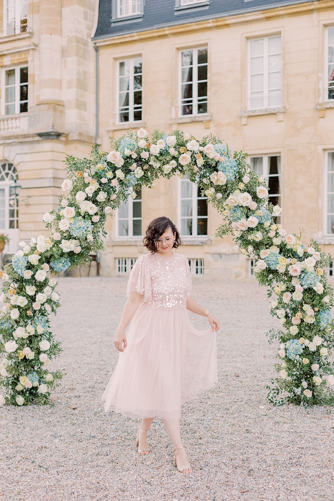 Frau mit braunen, lockigen Haaren läuft in Frankreich vor einem Chateau vor einem Blumenbogen. Sie trägt einen rosa Rock und ein rosa Paillettenshirt.