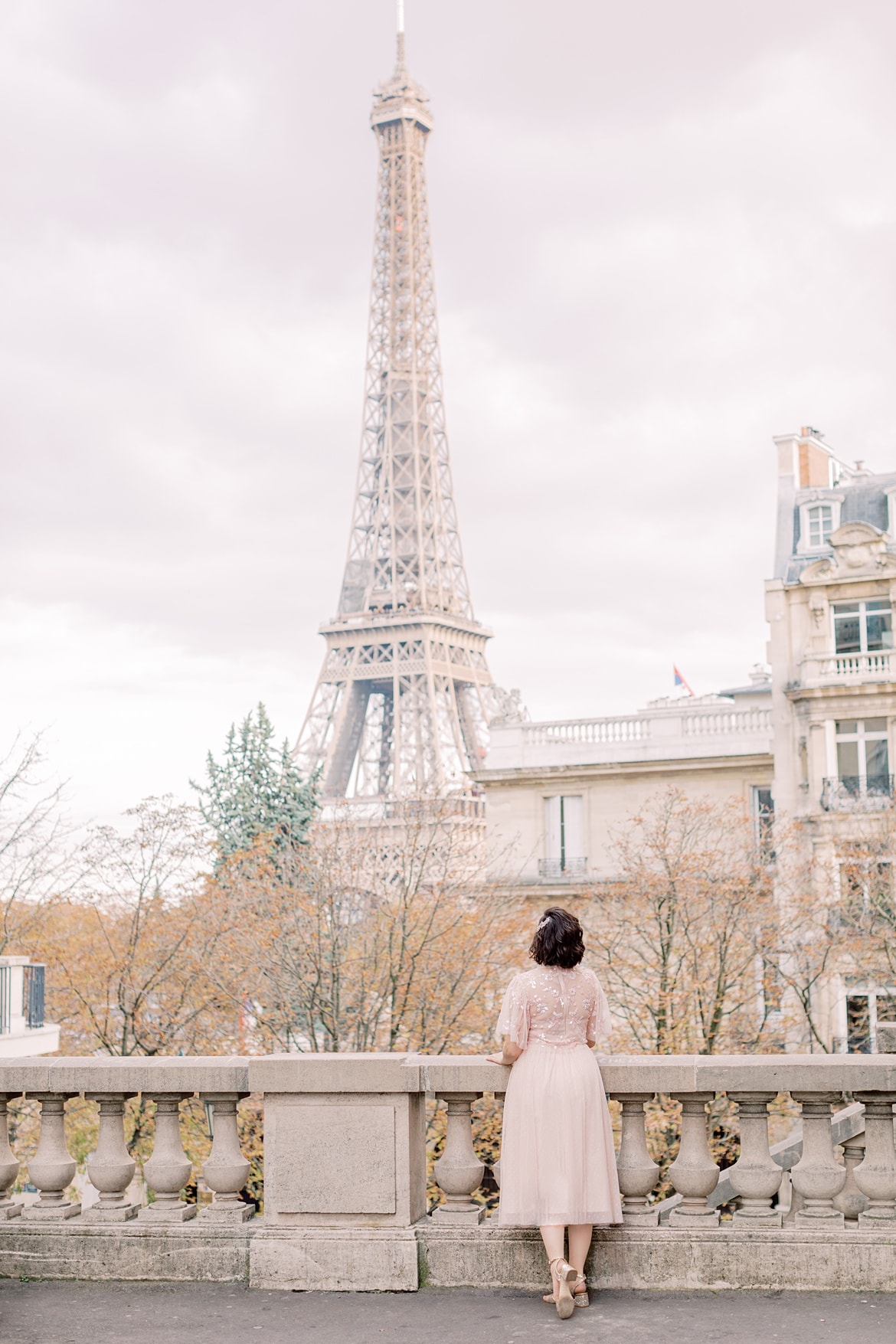 Frau mit braunen, lockigen Haaren steht in Paris auf einer Straße und schaut auf den Eiffelturm. Sie trägt einen rosa Rock und ein rosa Paillettenshirt.