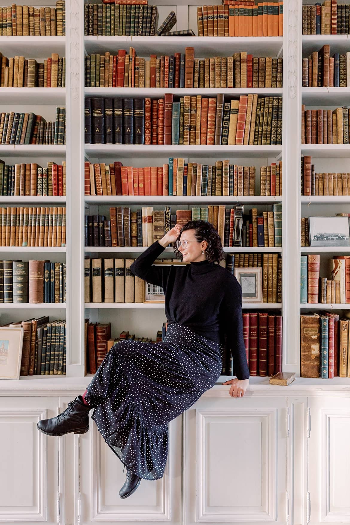 Frau mit dunklen, lockigen Haaren sitzt sehr vor einer Bücherwand in einer Bibliothek. Sie trägt ein schwarzes Kleid und einen schwarzen Pullover.
