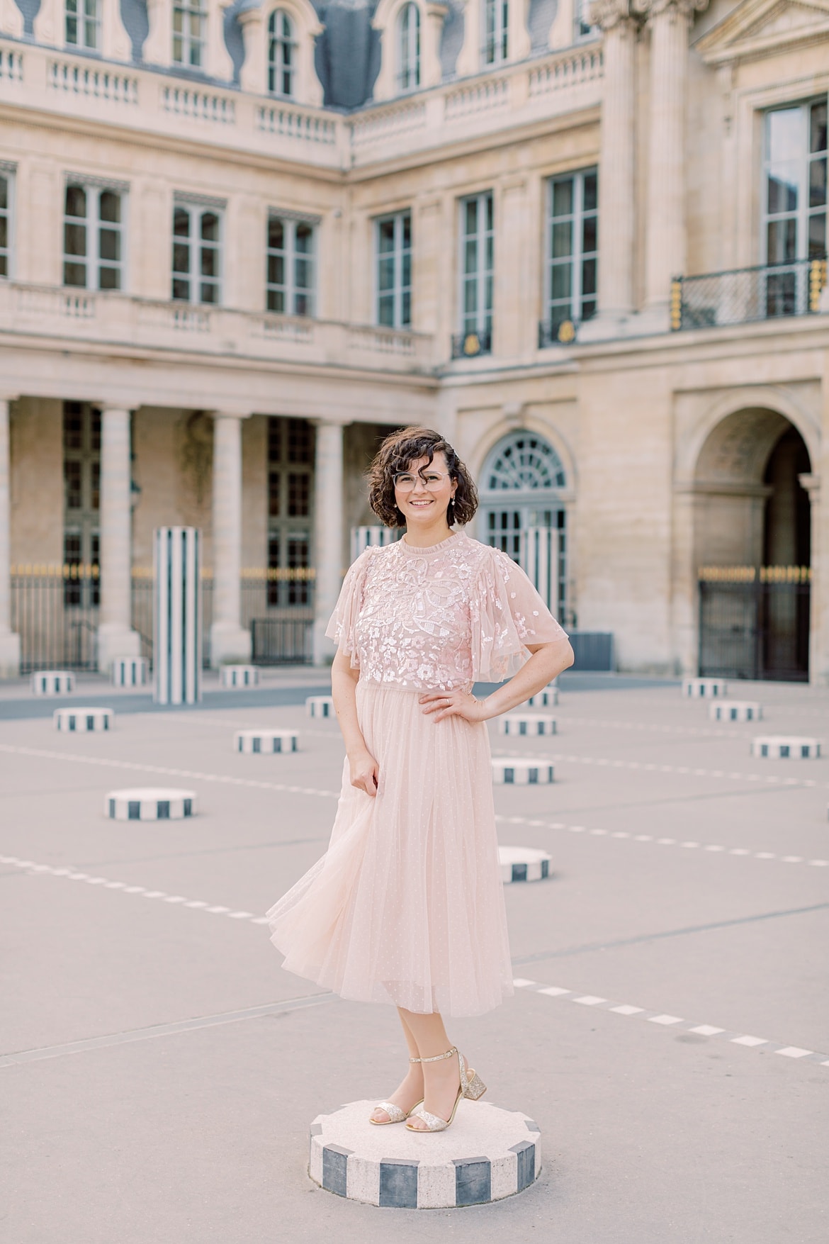 Frau mit braunen, lockigen Haaren lacht in die Kamera. Sie steht in Paris auf einer Säule im Palais Royal. Sie trägt einen rosa Rock und ein rosa Paillettenshirt.