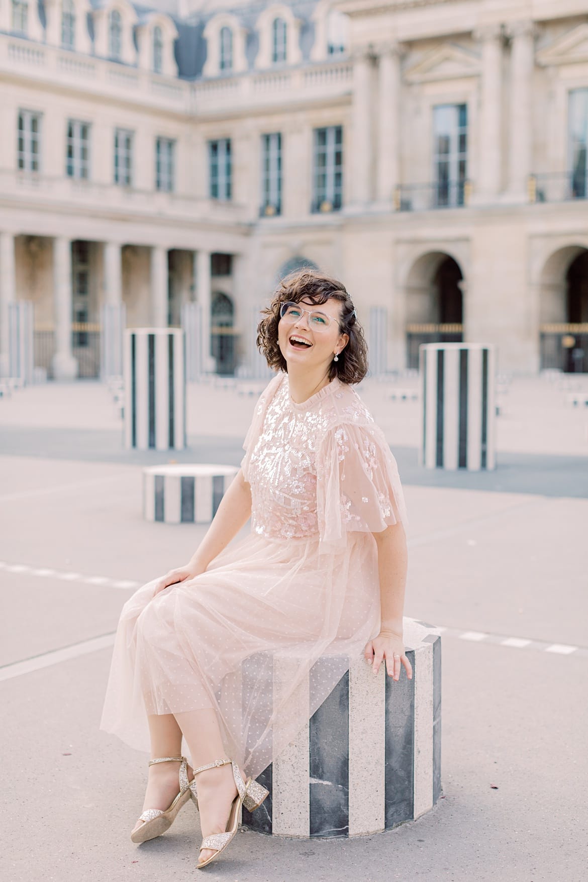 Frau mit braunen, lockigen Haaren lacht in die Kamera. Sie sitzt in Paris auf einer Säule im Palais Royal. Sie trägt einen rosa Rock und ein rosa Paillettenshirt.