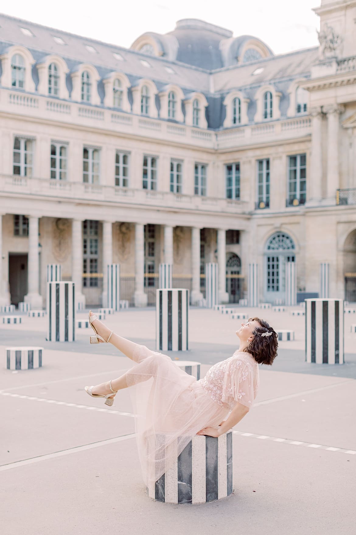 Frau mit braunen, lockigen Haaren sitzt in Paris auf einer Säule im Palais Royal. Man sieht sie von der Seite. Sie schaut nach oben zum Himmel. Sie trägt einen rosa Rock und ein rosa Paillettenshirt.