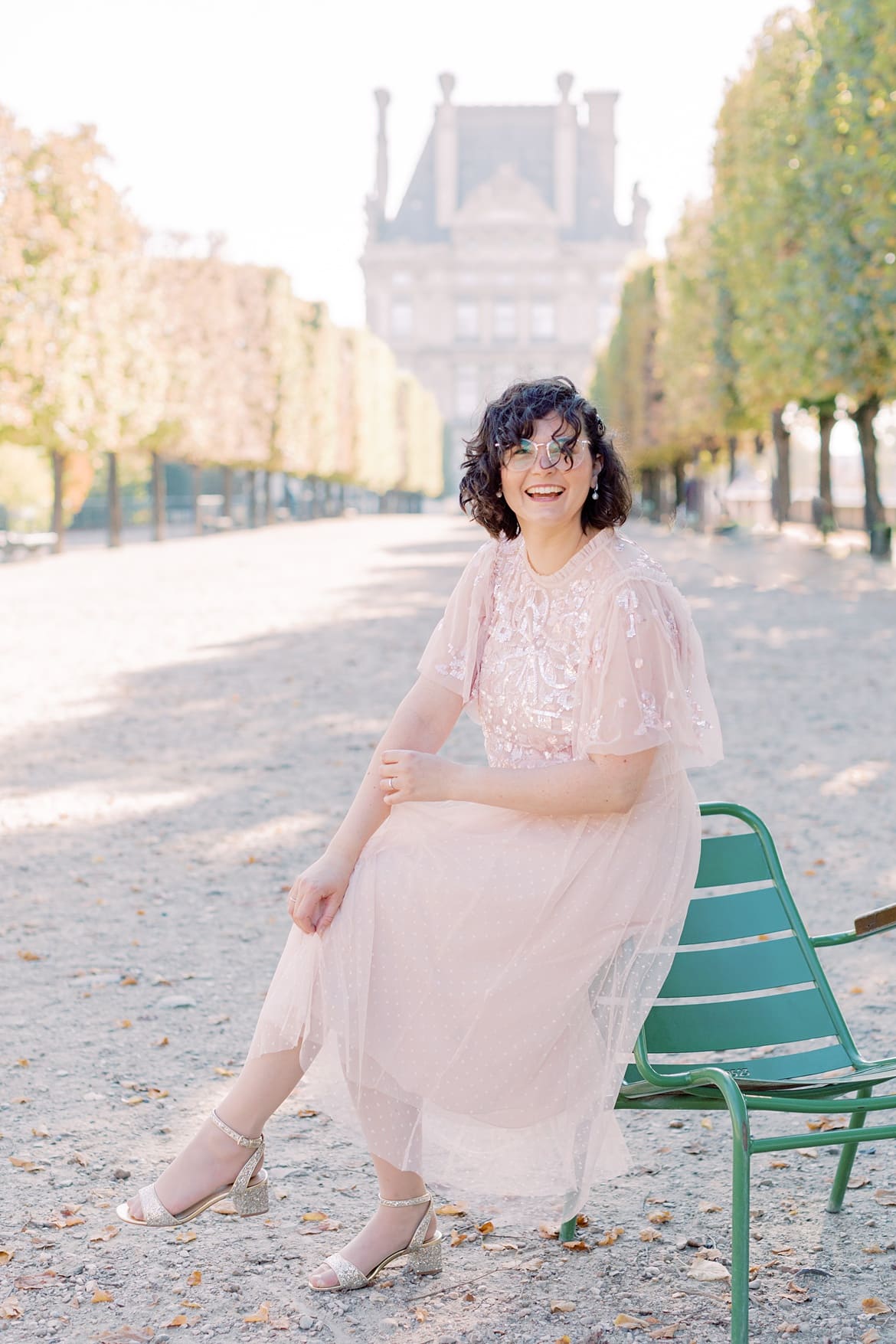 Frau mit braunen, lockigen Haaren lacht in die Kamera. Sie sitzt in Paris auf einem Stuhl im Jardin de Tuileries. Sie trägt einen rosa Rock und ein rosa Paillettenshirt.