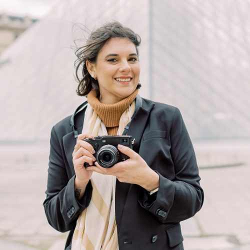 Eine Frau mit kurzen braunen Haaren, einem schwarzen Mantel und einem beigen Schal. Sie steht vor der Louvre Pyramide in Paris und hat eine Kamera in der Hand.